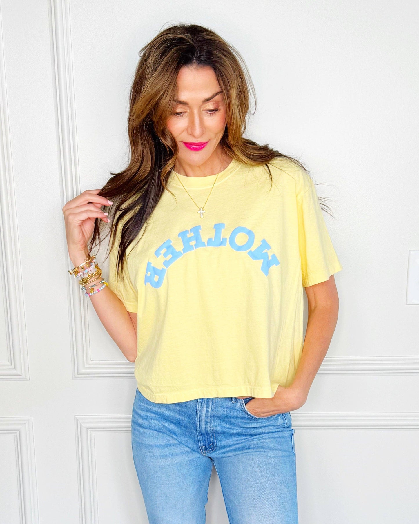 Woman wearing a yellow t-shirt with blue text and light blue jeans against a white wall.