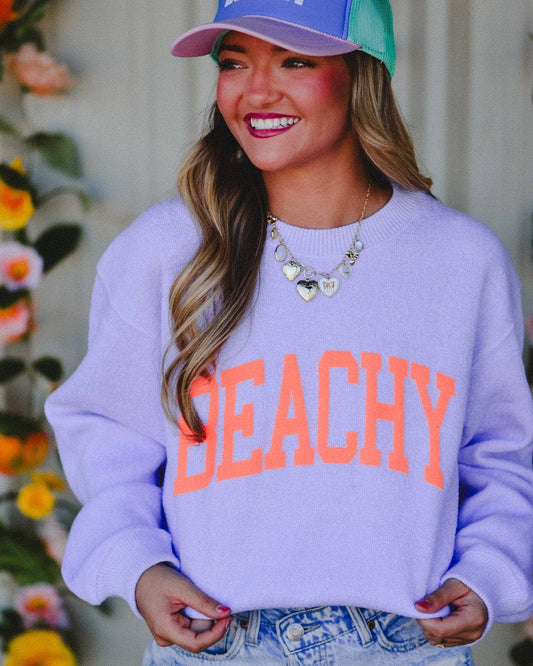 Woman wearing a purple 'BEACHY' sweatshirt and colorful cap, standing in front of floral decorations.