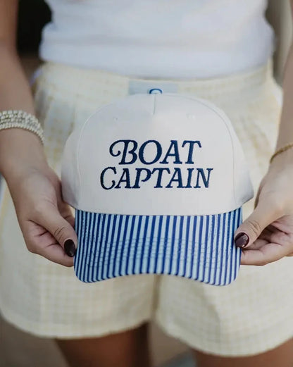 Cap with 'BOAT CAPTAIN' text held by a person wearing a white top and shorts.