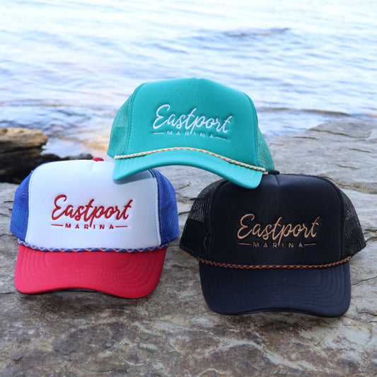 Three Eastport Marina hats in different colors on a rocky surface with water in the background