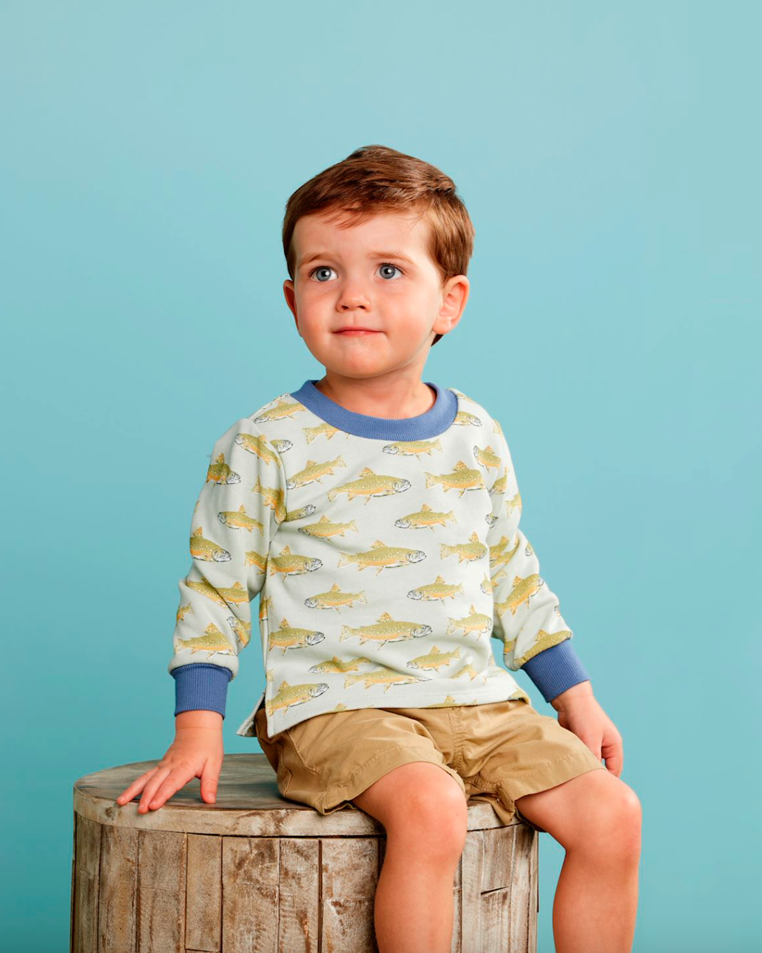 Young boy wearing a shirt with fish pattern sitting on a wooden block against a blue background
