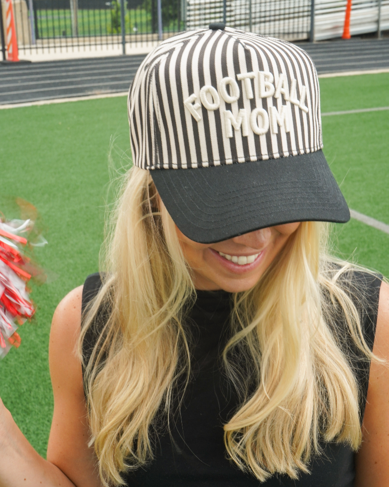 A woman on a sports field wearing a black and white striped baseball cap with 'FOOTBALL MOM' embroidered on the front, paired with a black sleeveless top.