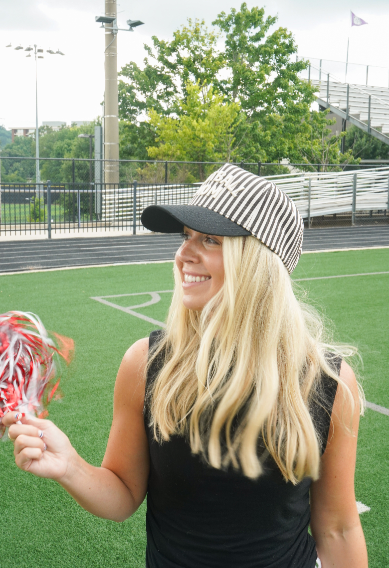 A smiling blonde woman on a sports field wearing a black and white striped baseball cap with a white script logo, a black sleeveless top, and holding red and white pom-poms.