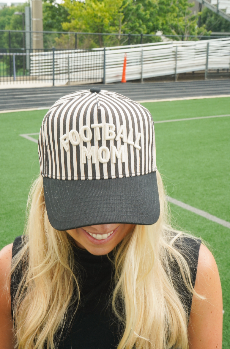 A woman on a sports field wearing a black and white vertically striped baseball cap with 'FOOTBALL MOM' embroidered in white across the front, with a black visor.