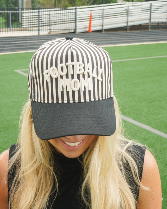 A woman on a sports field wearing a black and white vertically striped baseball cap with 'FOOTBALL MOM' embroidered in white across the front, with a black visor.