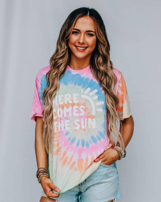 Woman wearing a tie-dye t-shirt with 'Here Comes the Sun' text against a plain background