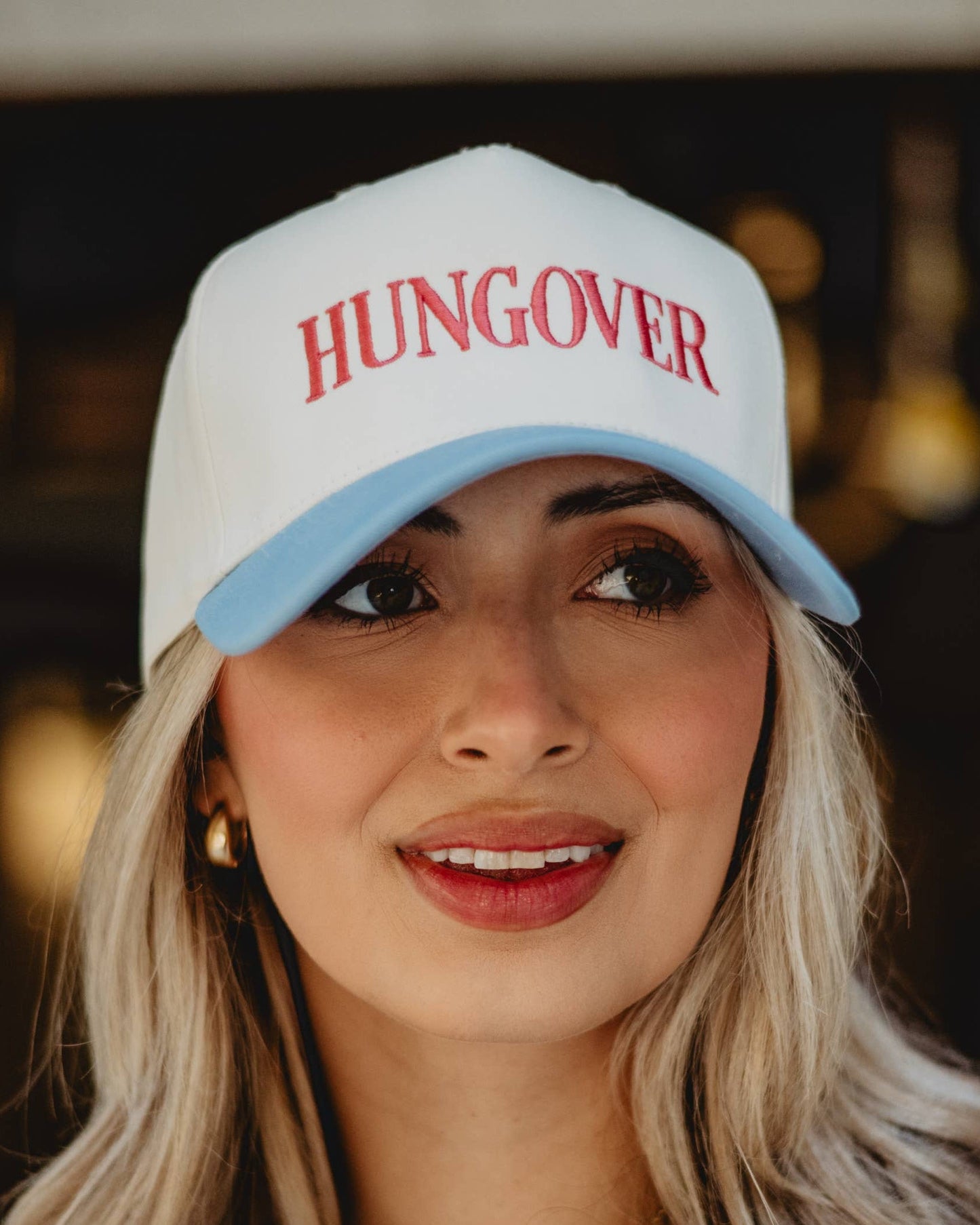 Woman wearing a white cap with 'HUNGOVER' text in red on a blurred background