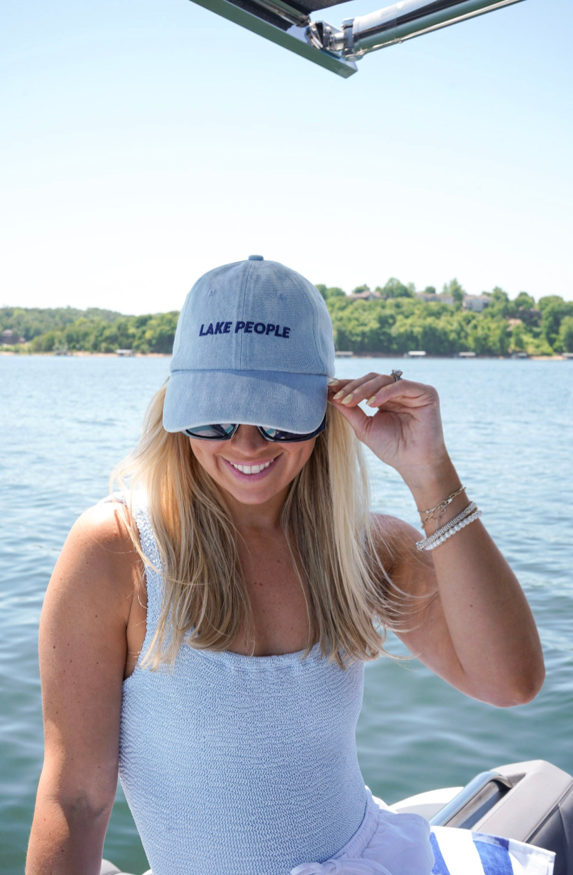 A woman on a boat wearing a light blue baseball cap with 'LAKE PEOPLE' embroidered in navy on the front, along with a matching light blue top.