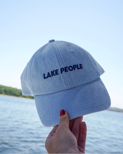 A light blue denim baseball cap with 'LAKE PEOPLE' embroidered in navy on the front, held by a hand against a background of water and sky.