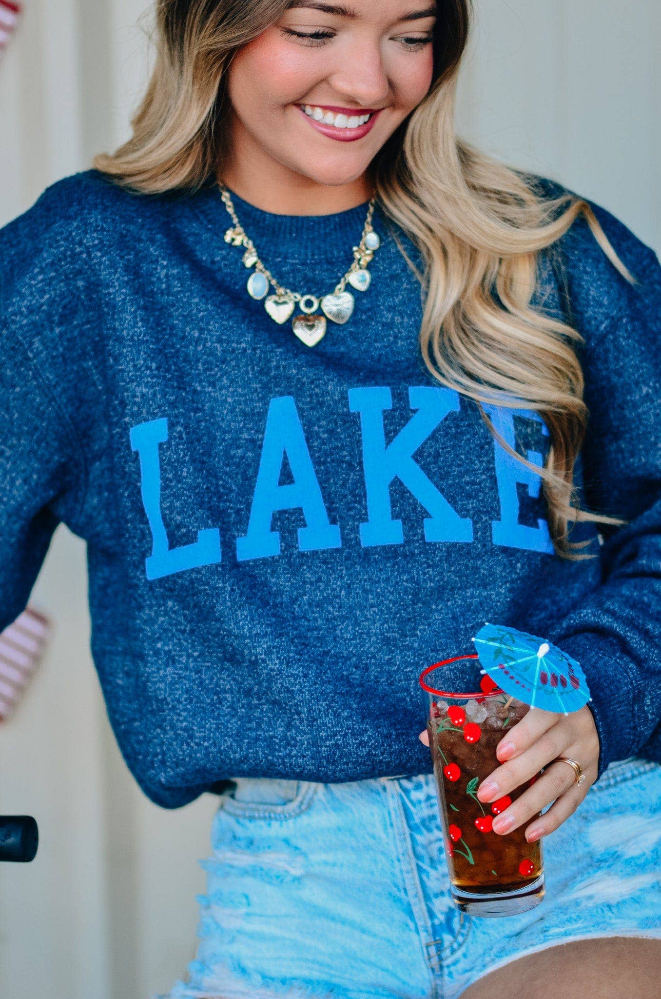 Woman wearing a blue 'LAKE' sweatshirt holding a drink.