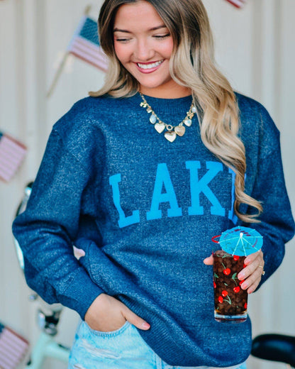 Woman wearing a blue sweatshirt with 'LAKE' text, holding a drink, standing in front of a bicycle and flags.