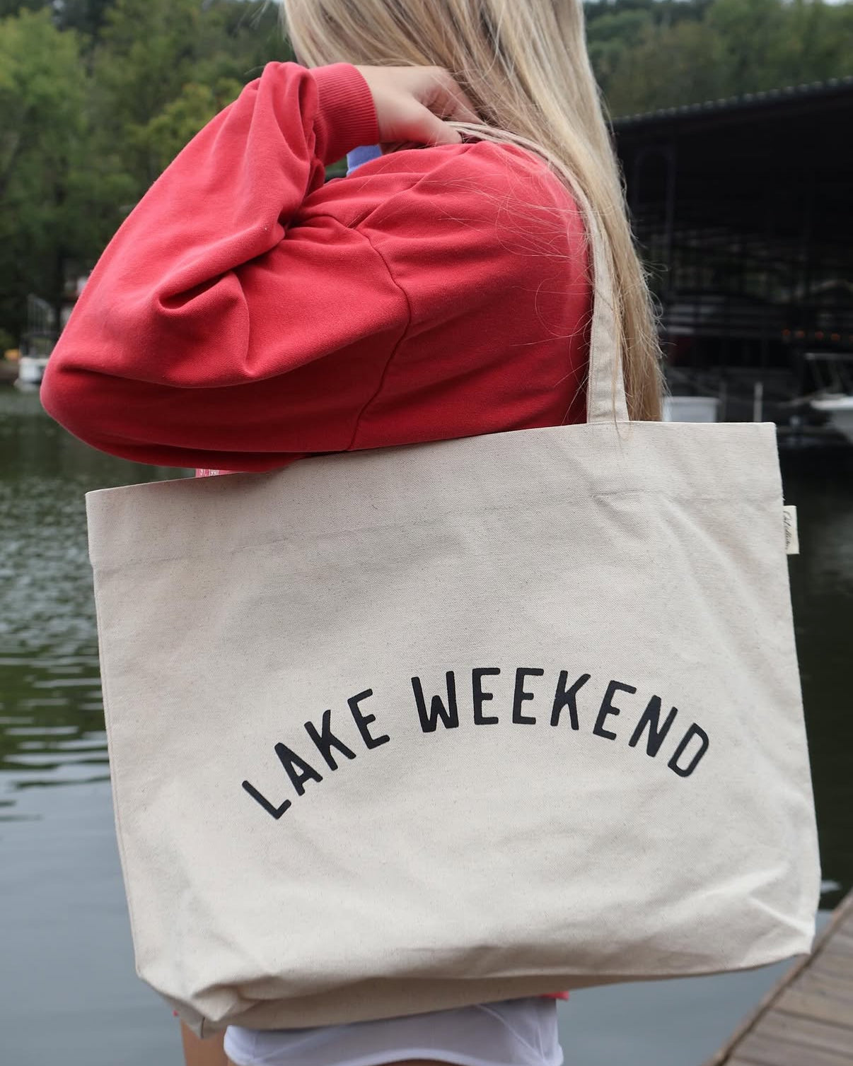 Person holding a tote bag with 'Lake Weekend' text by a lake.