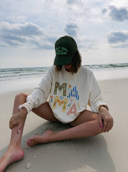 Person sitting on a beach wearing a white sweatshirt with colorful letters and a green cap.