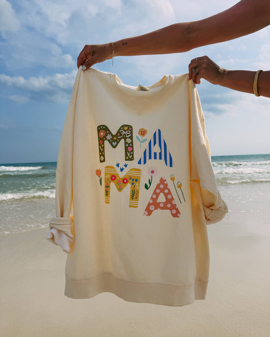 Beige sweatshirt with colorful letters spelling 'MAMA' held by a person on a beach.