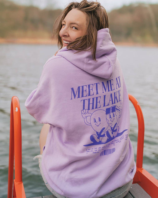 Person wearing a purple hoodie with 'Meet Me at the Lake' text and graphic, sitting on a dock by a lake.
