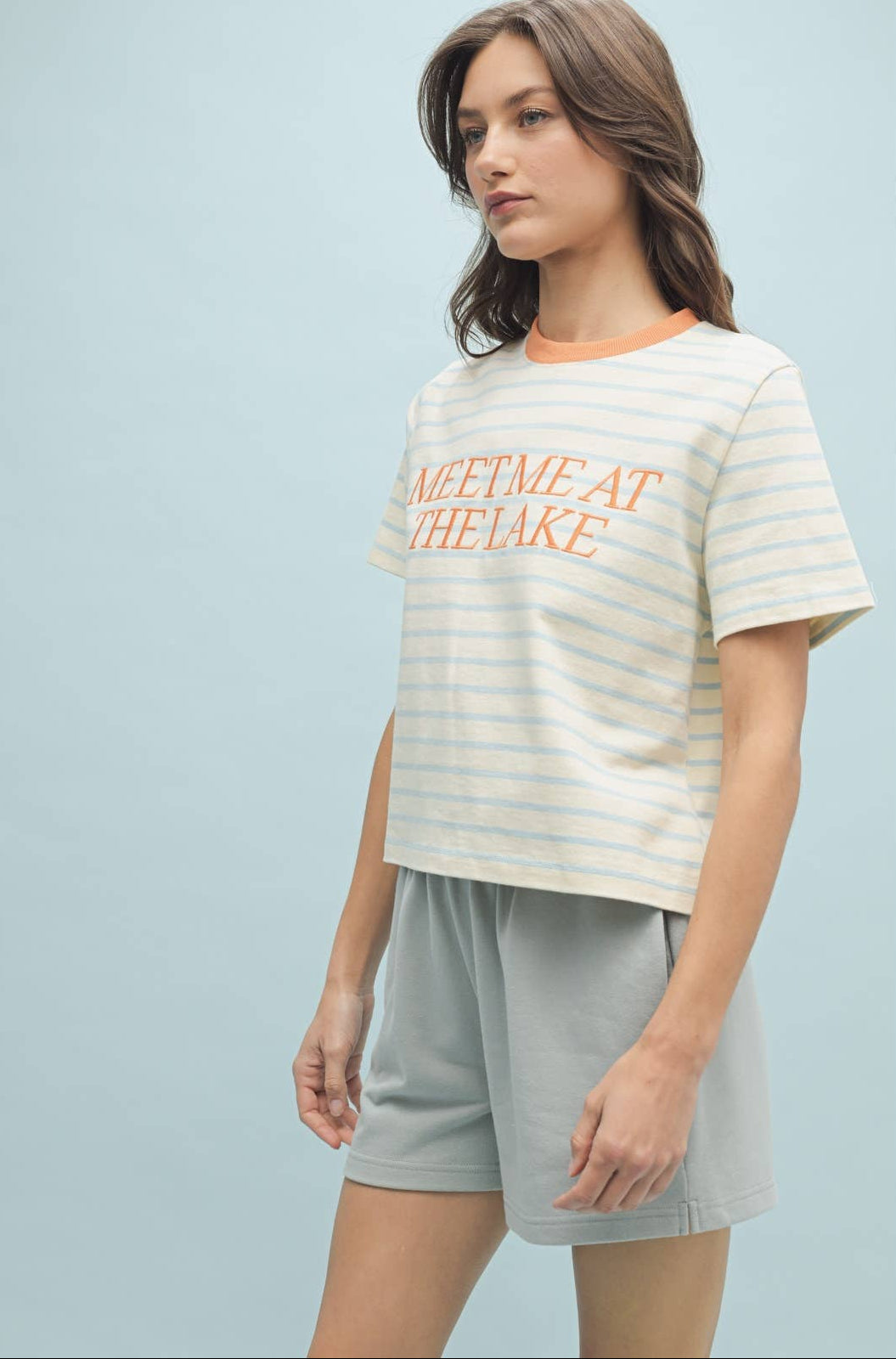 A woman wearing a striped knit top with the text 'MEET ME AT THE LAKE' on it, in a color palette that includes blue and white tones.