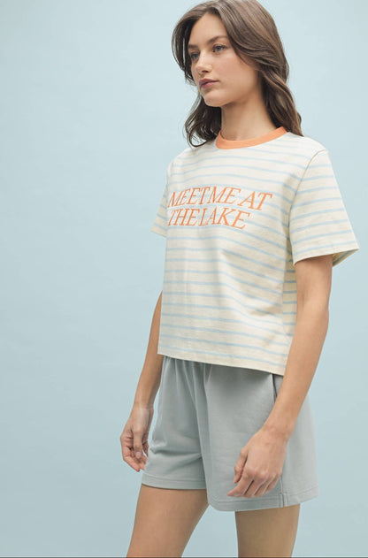 A woman wearing a striped knit top with the text 'MEET ME AT THE LAKE' on it, in a color palette that includes blue and white tones.