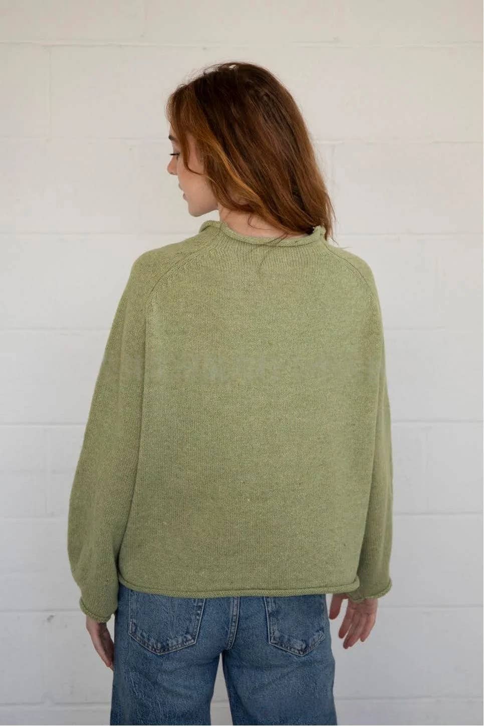 Person wearing a green sweater and blue jeans against a white wall