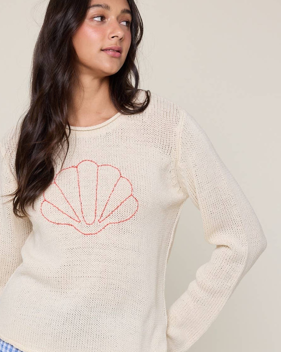 Woman wearing a cream sweater with a red shell design against a plain background