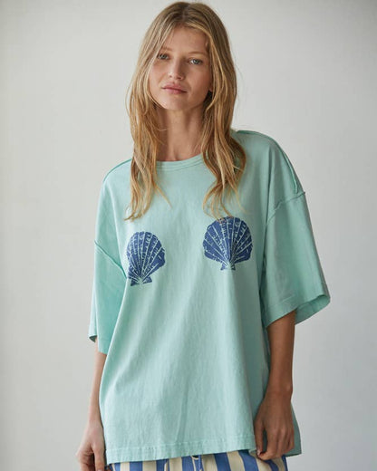 Woman wearing a light green oversized t-shirt with shell designs and striped shorts on a plain background