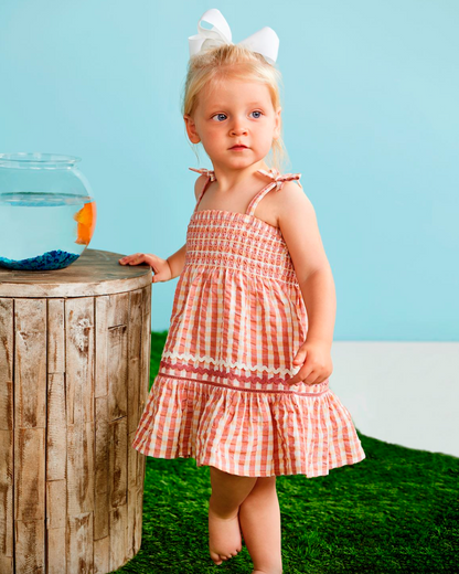 Young girl in a checkered dress standing next to a wooden post with a fishbowl against a blue sky.
