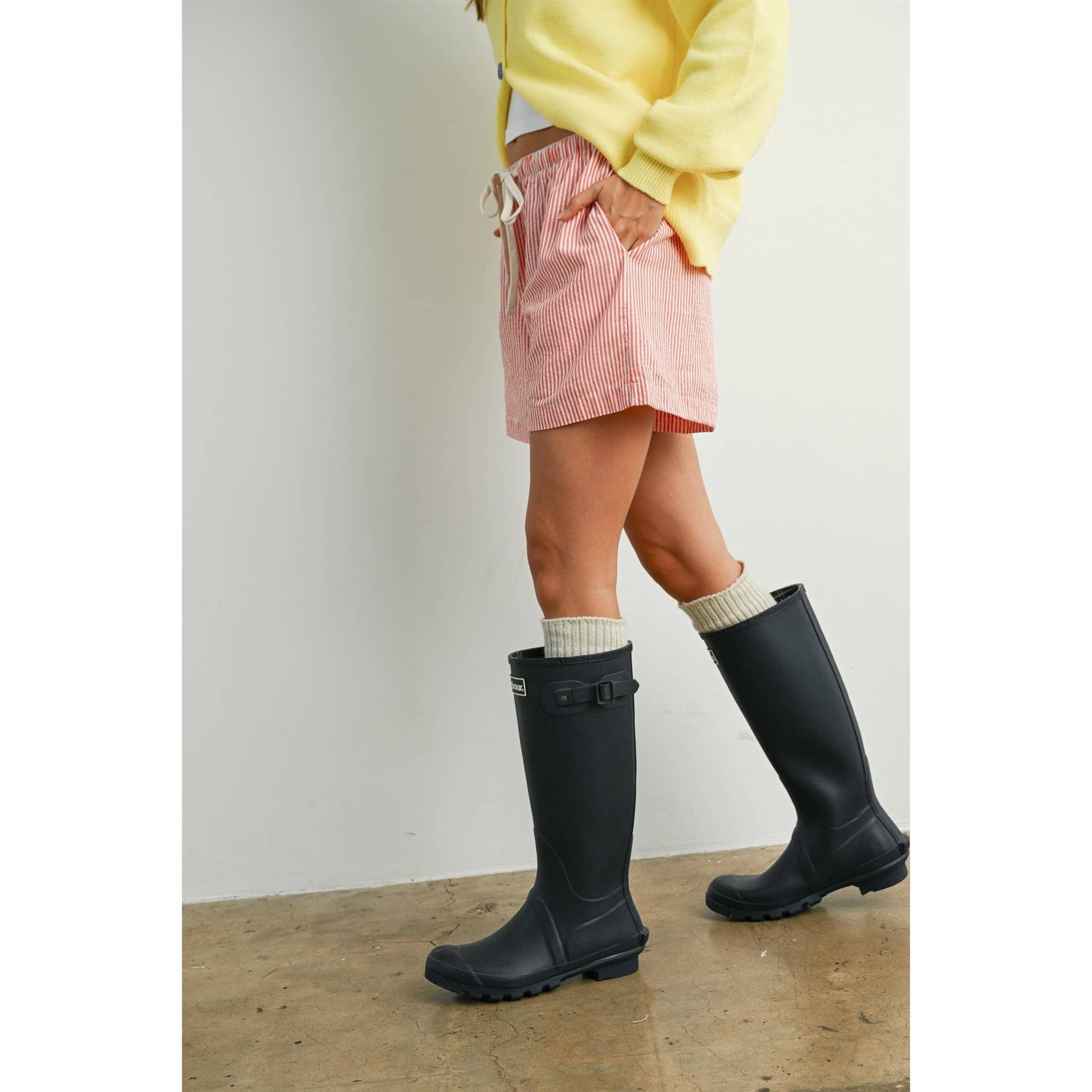 Person wearing black rain boots, a yellow sweater, and pink shorts on a wooden floor.