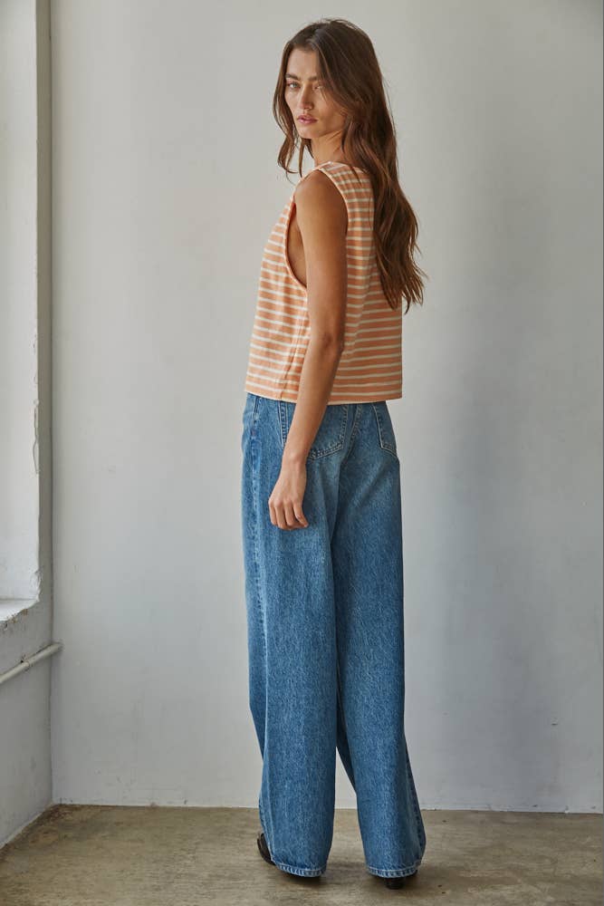 Woman wearing a striped tank top and blue jeans standing against a plain wall.