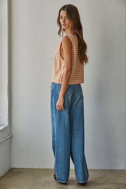 Woman wearing a striped tank top and blue jeans standing against a plain wall.
